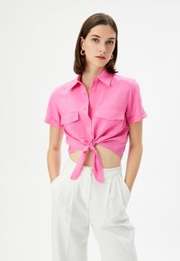 Camisa corta de manga corta en un brillante color rosa, con un nudo en la parte delantera y dos bolsillos en el pecho. Hecha de una tela texturizada, combinada con pantalones blancos.