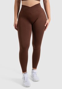 Bruine high-waisted leggings met een gestructureerde afwerking en een V-vormige tailleband, gecombineerd met witte sportschoenen.