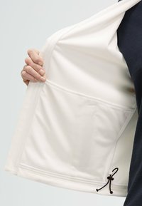 Giacca leggera con interno bianco, tasca in rete testurizzata e cordino regolabile all'orlo, con uno strato esterno scuro.