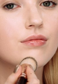 Make-up-Anwendung mit einem weichen, gerundeten Pinsel auf einem glatten Gesicht. Helle Hautfarbe, perlmuttartige Lippenfarbe und neutralfarbene Nägel sichtbar.