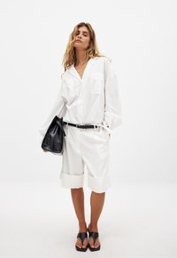 Weiße, übergroße Hemdbluse mit zwei Brusttaschen, kombiniert mit knielangen weißen Shorts, schwarzem Gürtel und schwarzen Schlipsandalen. Schwarze Handtasche.