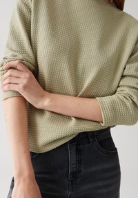 Donna che indossa una camicia a maniche lunghe verde chiaro con texture e maniche arrotolate, abbinata a jeans neri a vita alta, con una mano appoggiata sull'altro braccio.