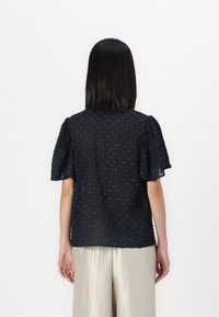 Top blu navy a maniche corte con tessuto texturizzato e dettagli ricamati delicati; vestibilità rilassata con un aspetto leggermente trasparente.