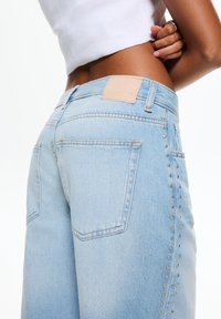 Jasnoniebieskie jeansy z denimu z jasnobrązową skórzaną naszywką, tylne kieszenie i srebrne ćwieki wzdłuż bocznego szwu. Konstrukcja dopasowana i o wyraźnym kroju.