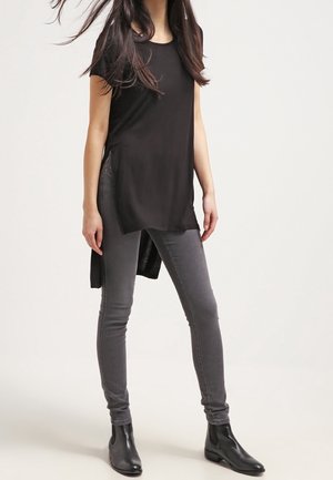 Femme debout portant un haut tunique noir à manches courtes, un jean skinny gris et des bottines noires sur un fond clair uni.