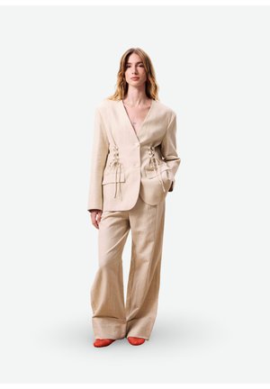 Femme portant un pantalon large beige et un blazer assorti avec des liens sur les côtés, associée à des chaussures rouges pointues, debout devant un fond blanc.