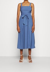 Mujer vestida con un vestido midi azul sin mangas con un lazo en la cintura y sandalias blancas de tacón alto, de pie frente a un fondo liso.