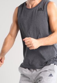 Muž v šedém bezrukávovém tričku Adidas a světle šedých šortkách Adidas v běžecké póze na jednolitém pozadí.