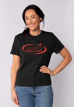 Femme souriante aux cheveux foncés portant un t-shirt noir avec le texte rouge « Beauty Babes Club » et un jean bleu, debout devant un fond clair uni.