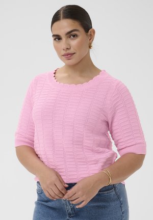 Kobieta o ciemnych włosach, nosząca złote kolczyki koła i bransoletkę, ubrana w różowy, teksturowany dzianinowy top oraz niebieskie dżinsy, pozująca na tle jednolitego tła.