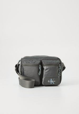 Sac à bandoulière en nylon gris avec deux poches zippées à l'avant, une bandoulière réglable et un accent logo. Texture lisse avec un design moderne.