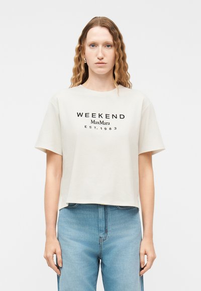 Γυναίκα με κυματιστά μαλλιά φοράει λευκό T-shirt με κοντό μανίκι και το κείμενο "WEEKEND MaxMara EST. 1983" μαζί με ανοιχτόχρωμο τζιν παντελόνι πάνω σε λευκό φόντο.