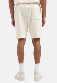 Pantaloni scurți albi texturați cu un model geometric, talie elastică și buzunar lateral, purtați cu adidași și șosete albe, văzuți din spate.