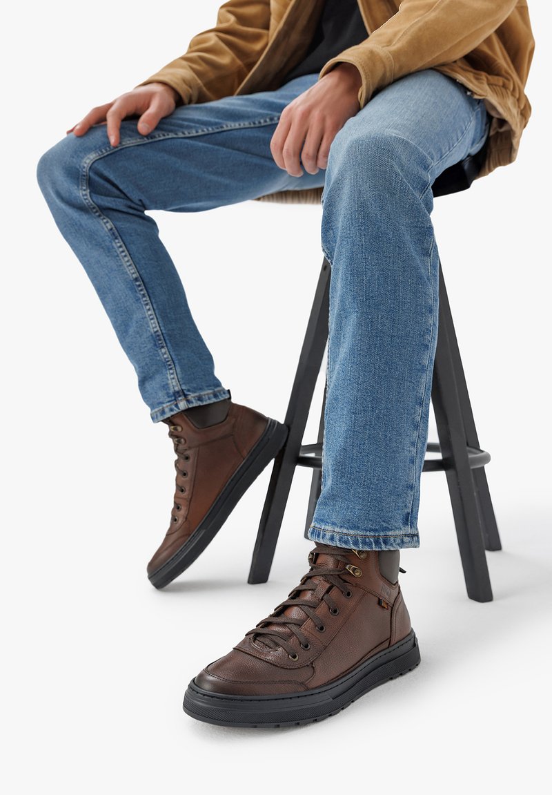 Rjave visoke športne copate iz teksturirane usnjene usnjene, okroglega nosu, zračnim gumijastim podplatom in kovinskimi obročki. Nosi se z modrimi jeans hlačami.