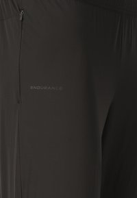 Nærbillede af sorte atletikbukser med en lodret lynlåslomme og "ENDURANCE"-logo trykt nær lommen.