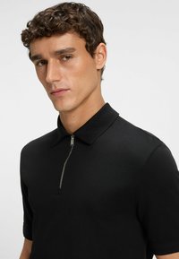 Czarna koszulka polo z krótkim rękawem i zamkiem przy kołnierzyku, wykonana z gładkiego, teksturowanego materiału. Charakteryzuje się dopasowanym krojem.