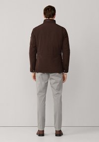 Mand iført en mørkebrun jakke, lysegrå bukser og brune sko, der står vendt mod en ensfarvet lysegrå væg.