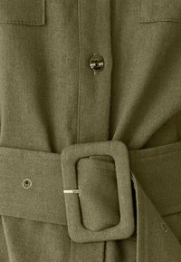 Tkanina olivno zelene barve z teksturirano površino, z gumbi spredaj, pasnim trakom s pravokotno sponko in enim žepom na prsih.