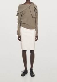 Pull beige à épaules dénudées avec détails drapés, associé à une jupe blanche en satin mi-longue et des chaussures noires à lacets.