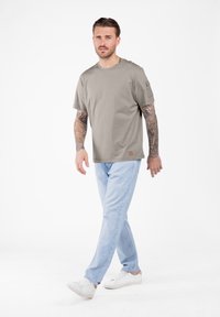 Hombre con los brazos completamente tatuados, vistiendo una camiseta gris claro, jeans de mezclilla azul claro y zapatillas blancas, caminando sobre fondo blanco.