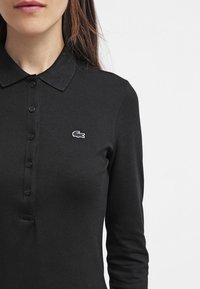 Polo noir à manches longues, avec une patte de boutonnage et un petit logo brodé blanc sur la poitrine gauche. Fabriqué en tissu texturé.