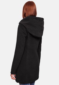 Black long coat with a large hood, made from textured fabric. Features subtle branding on the back and side slits for ease of movement.