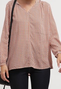 Blusa leggera a maniche lunghe in arancione e bianco con un motivo geometrico, scollo a V e una texture morbida, abbinata a jeans scuri.