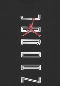 Černé tričko s červeným logem Jordan a bílým textem "JORDAN" vertikálně zarovnaným v tučném sportovním písmu.