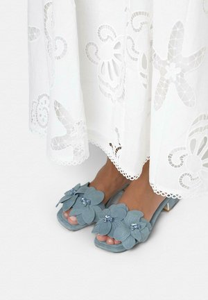 Sandali blu con tacco decorati con grandi fiori e centri gioiello, indossati con un vestito bianco caratterizzato da motivi ricamati traforati.