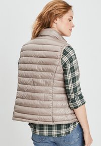 Beige Steppweste mit horizontalem Quilt-Design, hohem Kragen und lockerer Passform. Über einem grün-weißen Karomusterhemd mit aufgerollten Ärmeln getragen.
