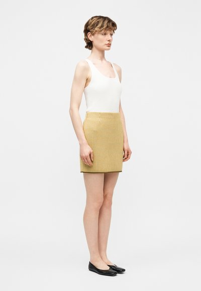 Νεαρή γυναίκα που φοράει λευκό αμάνικο τοπ, κίτρινη φουστα με καρό σχέδιο και μαύρα φλατ παπούτσια, στέκεται μπροστά σε απλό φωτεινό φόντο.