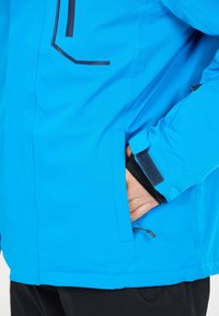 Blå vattentålig jacka med sidoficka, justerbara ärmslut och en mjuk yta, designad för utomhusaktiviteter. Svarta accenter framhävs.