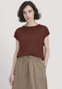 Frau mit kurzen lockigen Haaren, die ein braunes kurzärmeliges Oberteil trägt, das in beige Bundhose mit Kordelzug gesteckt ist, und gegen einen schlichten Hintergrund steht.