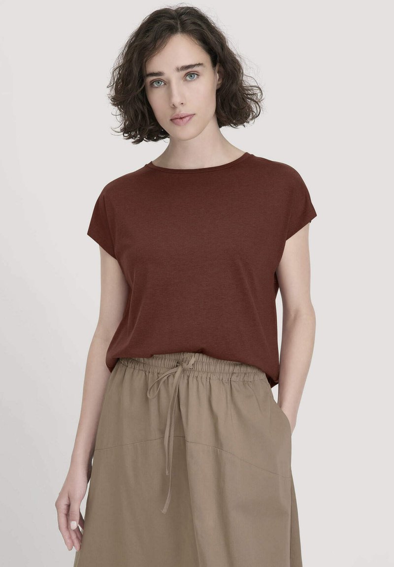 Frau mit kurzen lockigen Haaren, die ein braunes kurzärmeliges Oberteil trägt, das in beige Bundhose mit Kordelzug gesteckt ist, und gegen einen schlichten Hintergrund steht.