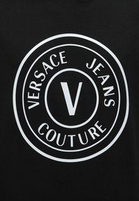 Μαύρο ύφασμα με λευκό κυκλικό λογότυπο Versace Jeans Couture που εμφανίζει ένα μεγάλο «V» στο κέντρο.