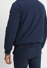 Man som bär en mörkblå bomberjacka och matchande byxor, sedd bakifrån med vänster hand avslappnat vid sidan.