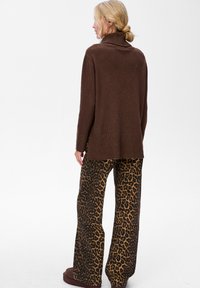 Kobieta z blond włosami związanymi w niski kok, nosząca złote kolczyki w kształcie kół, brązowy sweter z golfem i szerokie spodnie w panterkę, odwrócona plecami.