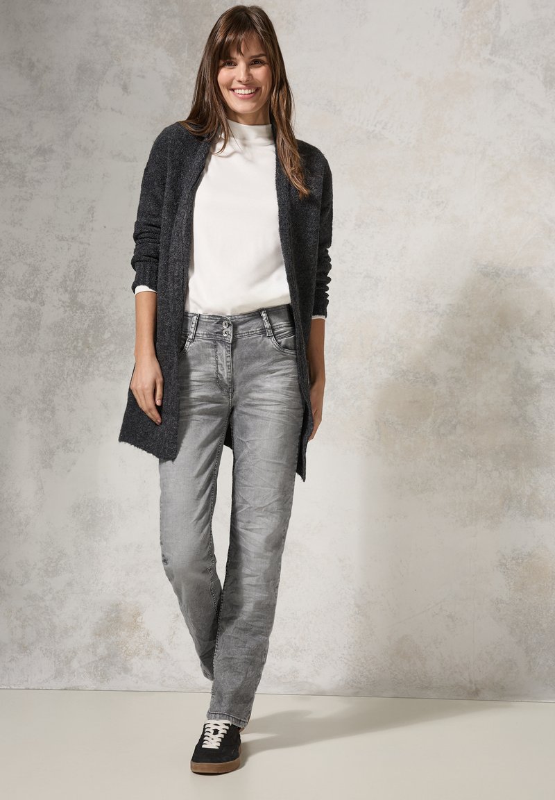 Long gilet gris foncé sur un col roulé blanc, associé à un jean gris clair et des baskets noires. Texture douce et coupe décontractée.