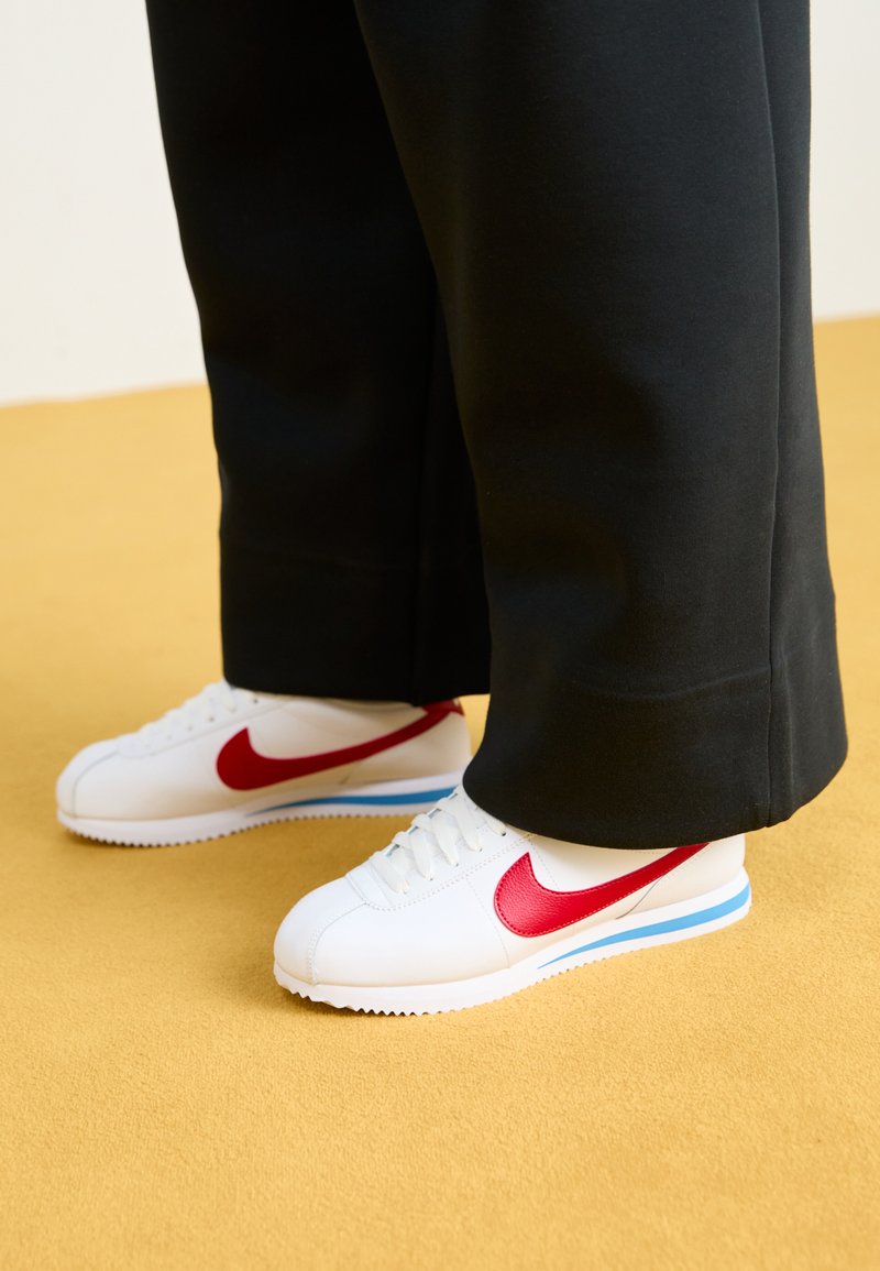 Hvide sneakers med rød Nike swoosh, tekstureret overdel og blå detalje på sålen, båret med sorte løstsiddende bukser på en gul baggrund.
