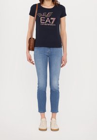 Femme portant un t-shirt bleu marine EA7 Emporio Armani, un jean bleu clair, des baskets beiges et un sac à bandoulière marron, debout sur un fond blanc.