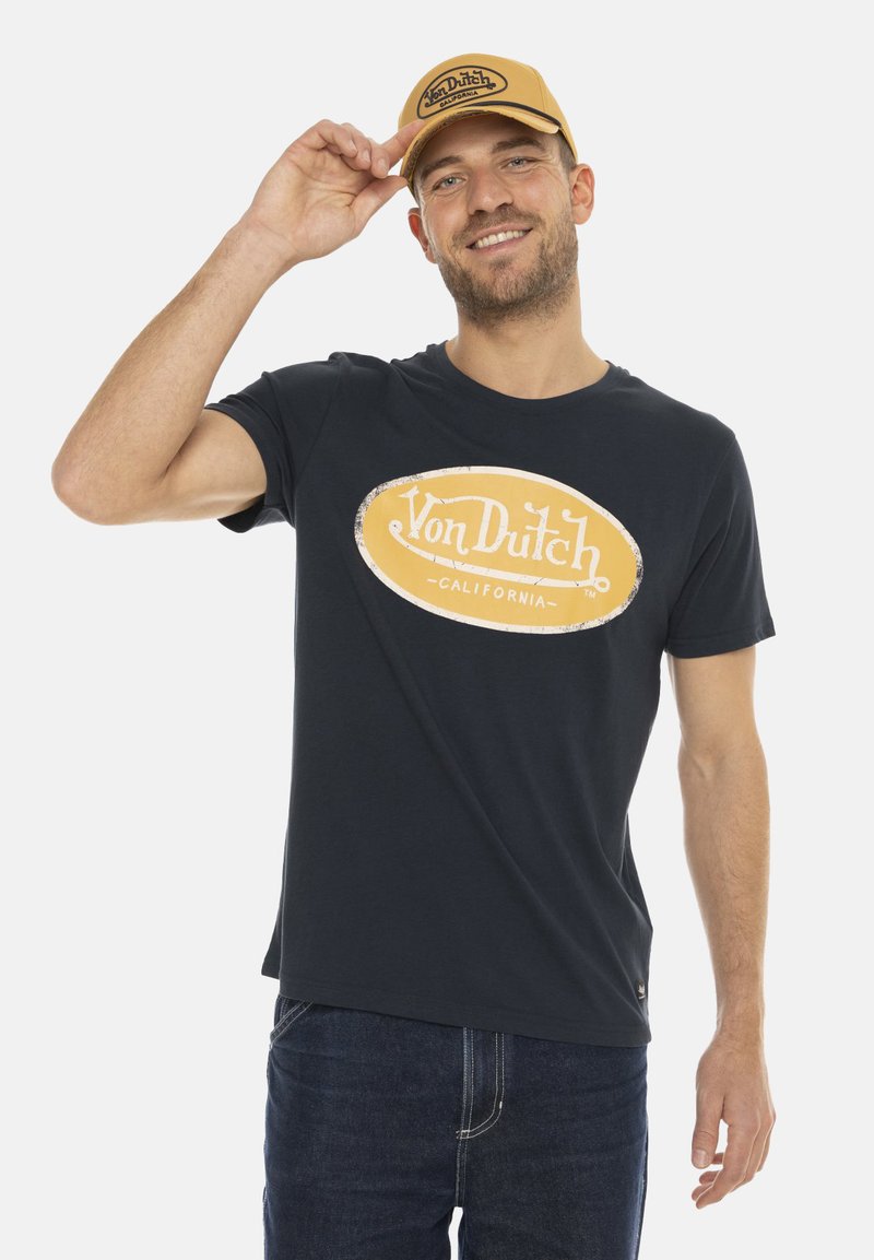 T-shirt marine avec un logo arrondi Von Dutch en jaune. Le modèle porte une casquette de baseball jaune et un jean bleu foncé, souriant tout en ajustant la casquette.