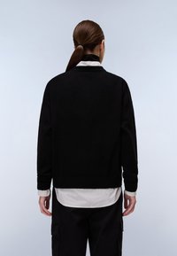 Maglione nero con una vestibilità rilassata, polsini e orlo a coste, indossato sopra una camicia a righe bianca e grigia. Lo sfondo è azzurro chiaro.
