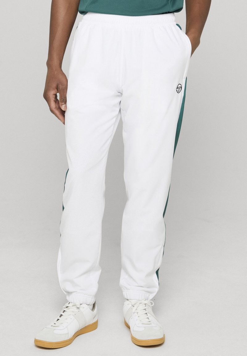 Férfi fehér melegítőnadrágban, zöld oldalsávval, rugalmas derék- és szegélyrésszel, fehér sportcipőben, amelynek gumitalpa van.