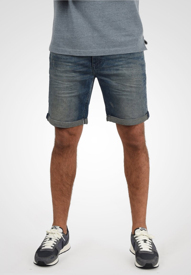 Shorts en denim d'une couleur bleu délavé, avec des revers roulés. Associés à des baskets grises et un t-shirt gris clair. La texture semble lisse.