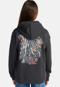 Persoon in een zwart hoodie met een afbeelding van twee snauwende, aan elkaar gebonden honden en een kleine rennende figuur, tekst "ELEMENT & TIMBER!" op de achterkant.