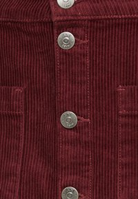 Chaqueta de pana burdeos con botones a presión plateados, textura de canalé vertical y dos bolsillos frontales para un detalle adicional.