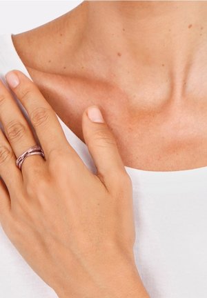 Main avec une bague en argent tressé reposant sur une clavicule nue, portant un haut blanc.