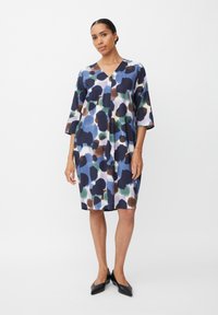 Eine Frau steht in einem lockeren, knielangen Kleid mit blauen, grünen, braunen und weißen abstrakten Flecken und schwarzen spitzen Ballerinas vor einem weißen Hintergrund.