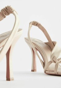 Beige High Heels Sandalen mit Slingback-Design, ausgestattet mit gerafften Riemen und glatter Leder-Oberfläche; offen an der Spitze und mit schlankem Absatz.