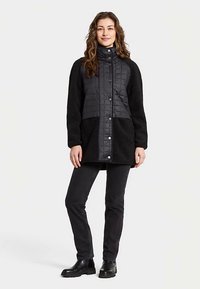 Zwarte gewatteerde donzen jas met zachte mouwen, hoge kraag en drukknopen, gecombineerd met zwarte broek en stevige zwarte laarzen.